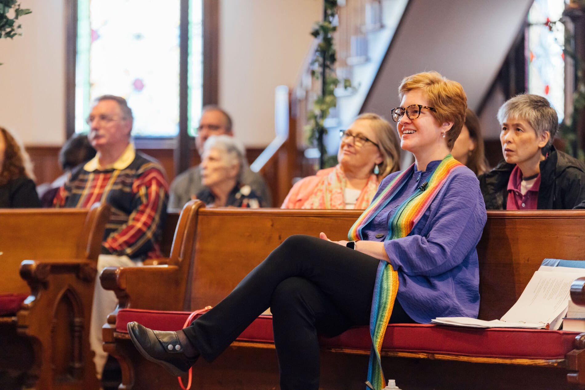 A woman wearing a rainbow stole sitting in a pew and smiling