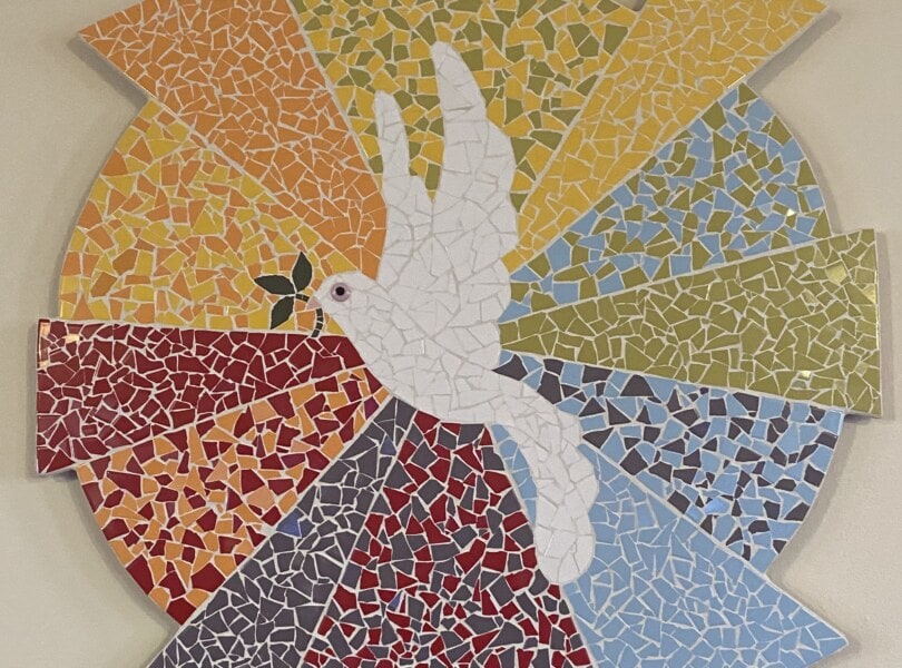 Mosiac artwork of a white dove holding an olive branch flying in front of a multicolored sun.