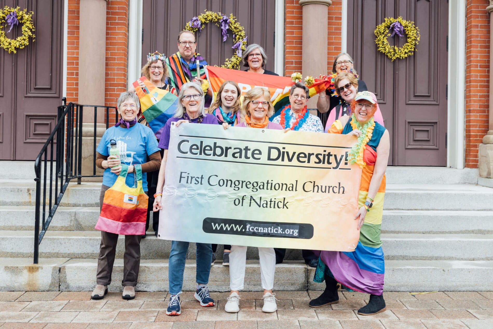 A group of people in front of a church wearing rainbow attire and holding a sign that says &quot;Celebrate Diversity - First Congregational Church of Natick&quot;