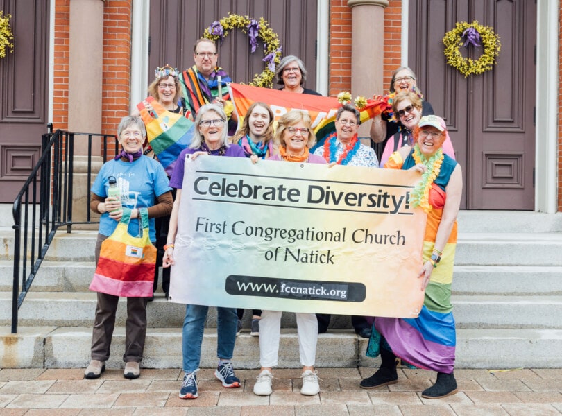A group of people in front of a church wearing rainbow attire and holding a sign that says "Celebrate Diversity - First Congregational Church of Natick"