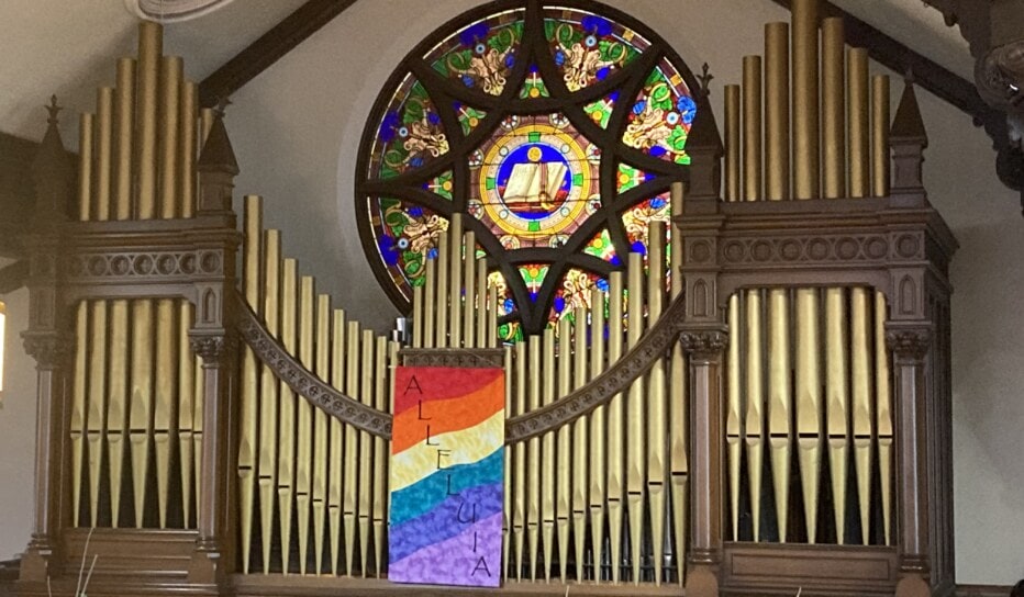 A large pipe organ with a rainbow banner draped over it and a stained glass window behind it.