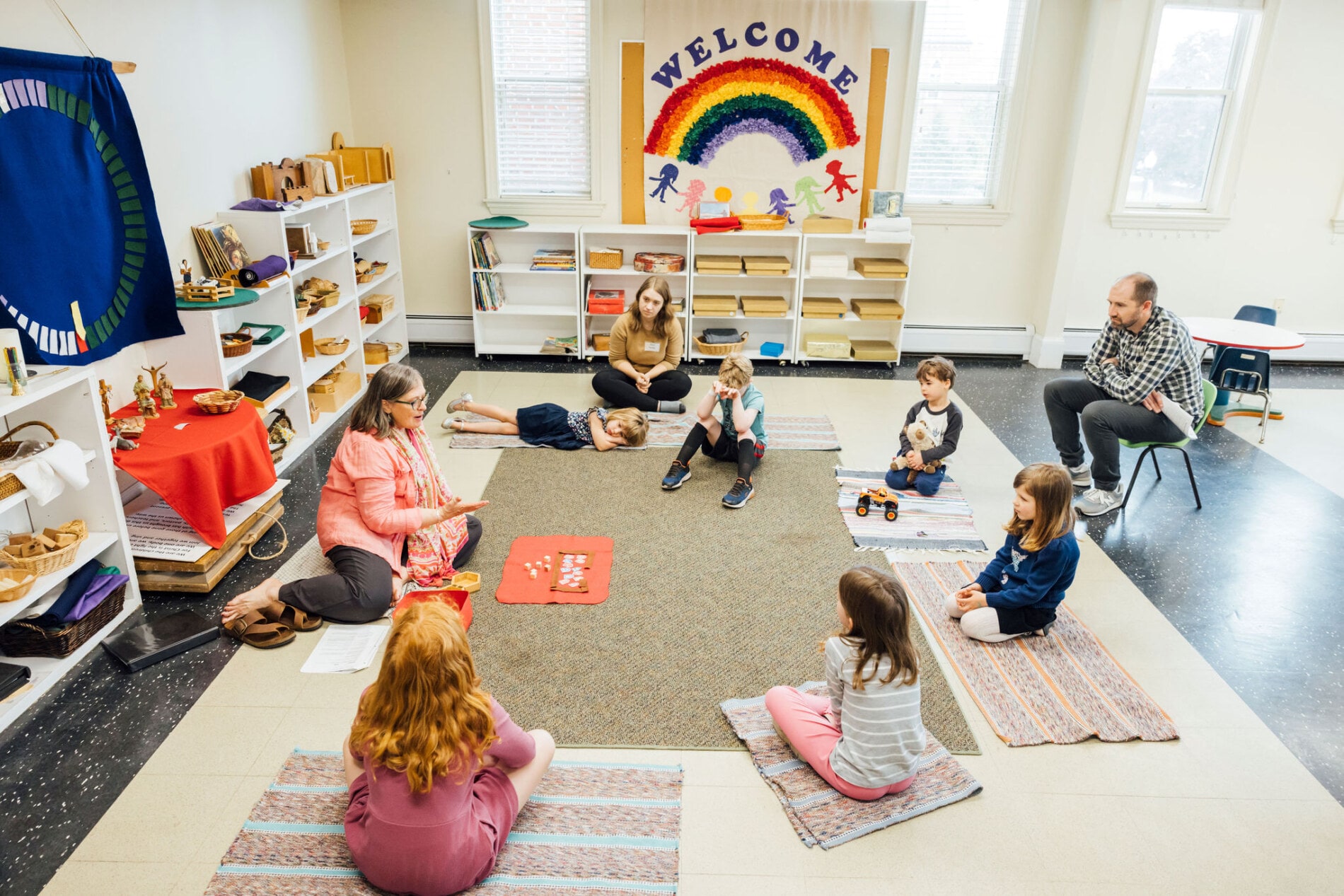 Two adults teach a group of children sitting in a circle on the floor