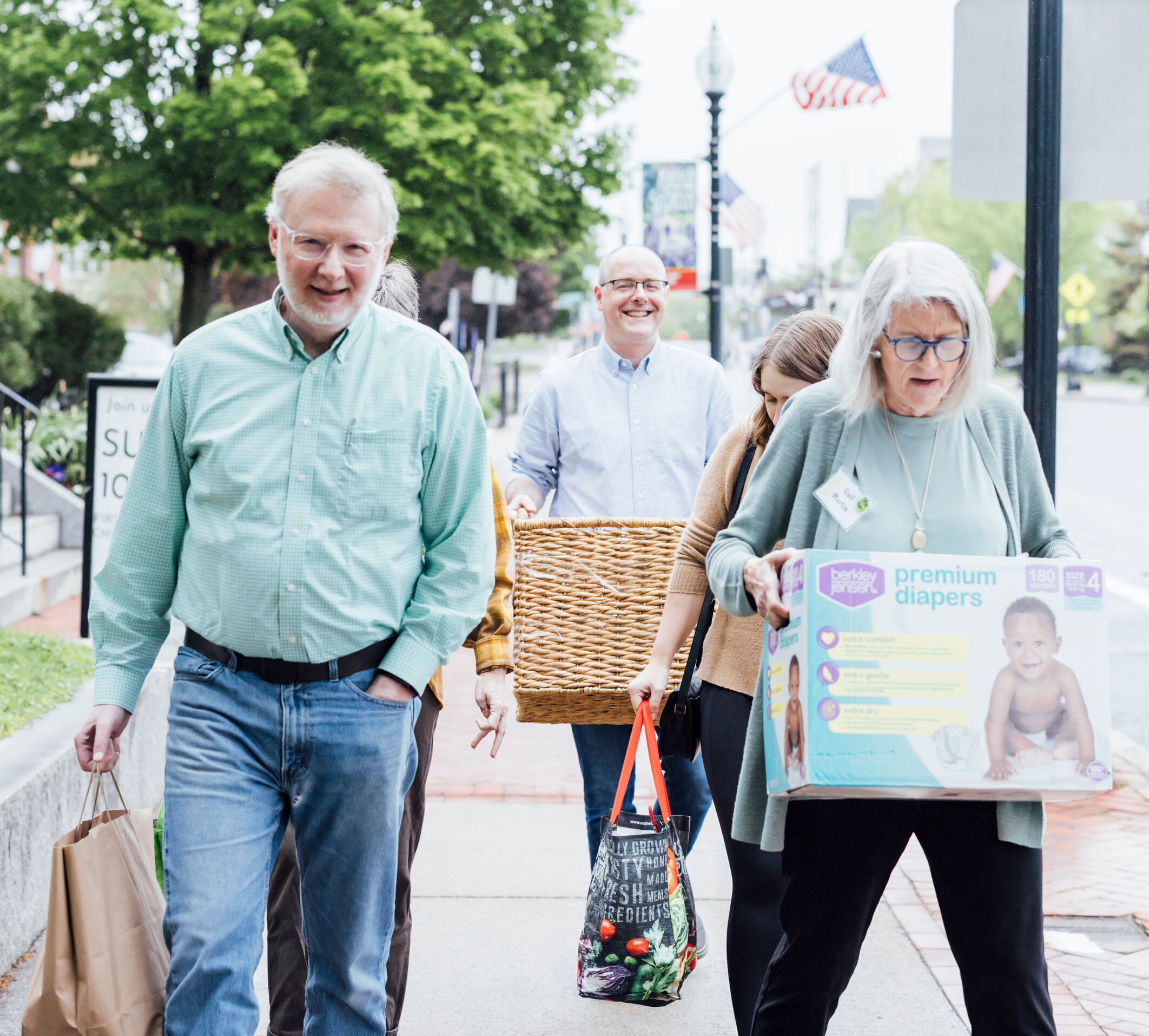 Pantry Parade Four people walking down a sidewalk carrying containers with food and diapers.