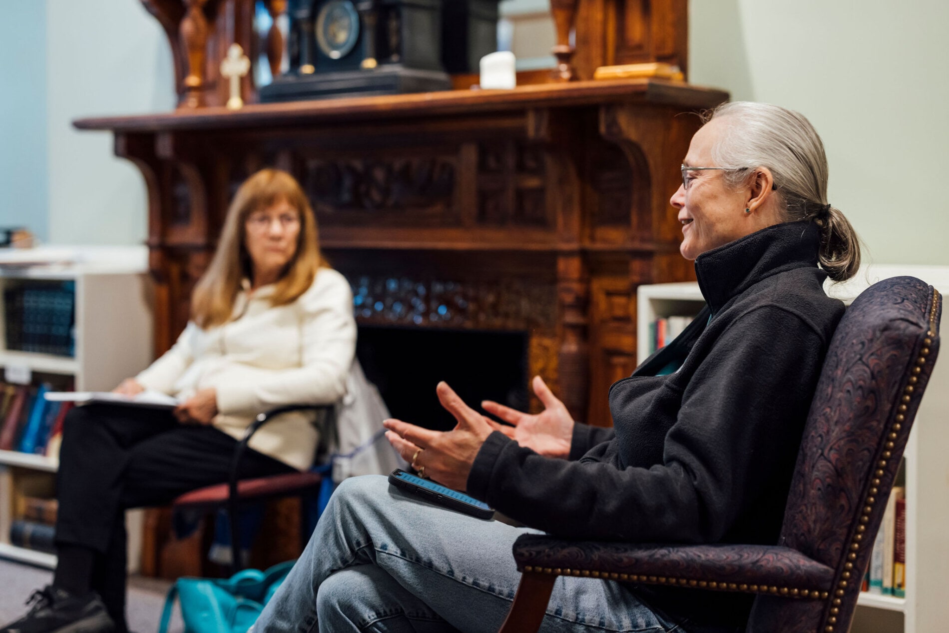 Two adults seated in chairs with a wooden fireplace mantel in the background