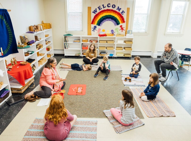 A circle of children sit on the floor while listening to an adult teacher.