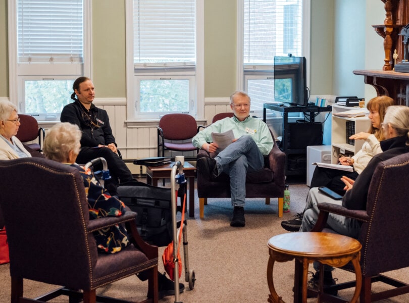 A circle of six adults sit in chairs while having a friendly discussion.