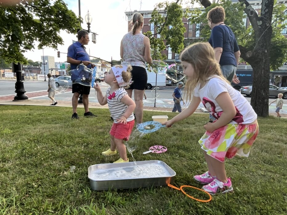 Children blow giant soap bubbles on church lawn.
