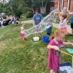 Children blow giant soap bubbles on church lawn.