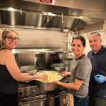 Three people putting a large pot of peeled potatoes on the stove