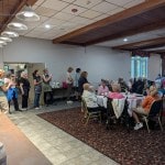 A group of volunteers watches as a room of people eat the meal their prepared.
