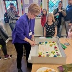 A women cuts a large cake as children and adults look on.